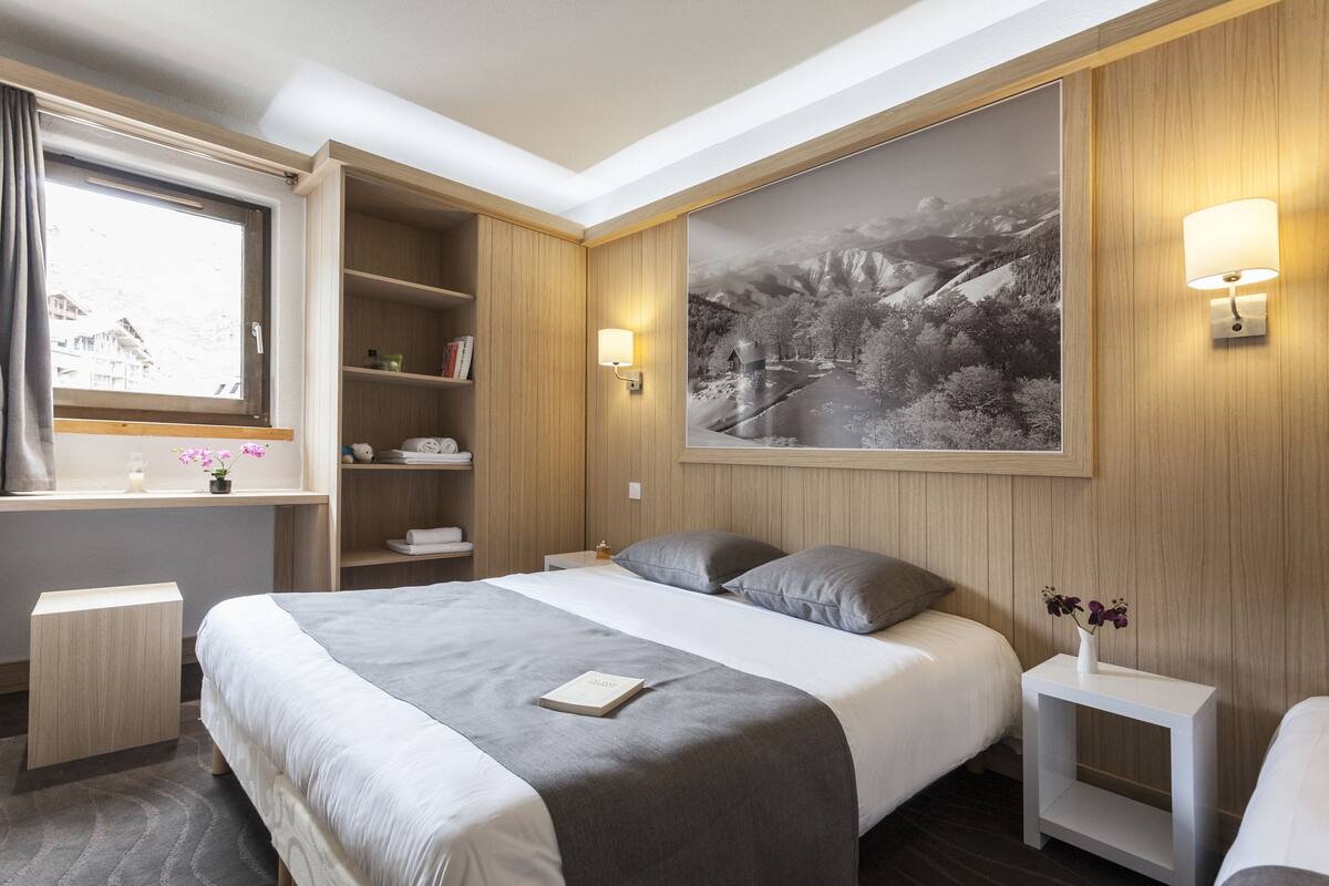 Una habitación de hotel moderna con una cama grande, mesitas de noche, una ventana con cortinas, una estantería y un cuadro grande en la pared.