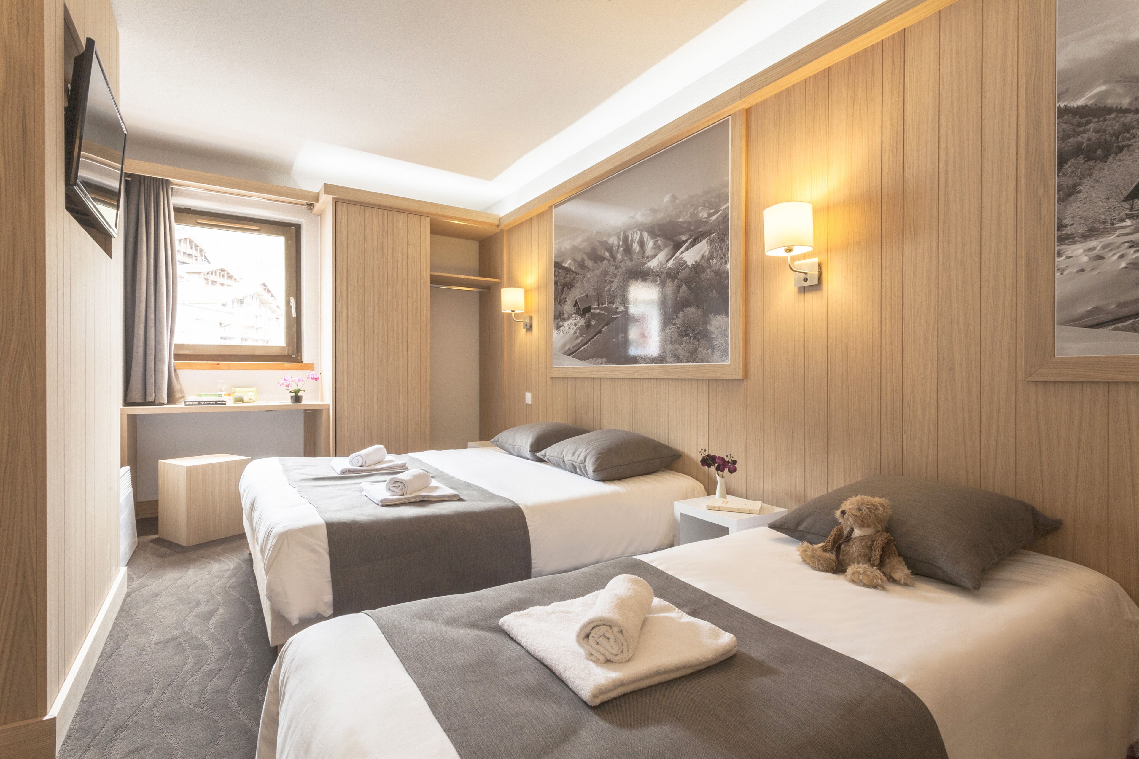 A hotel room with two beds, a large window, a flat-screen TV, and a wall-mounted lamp.