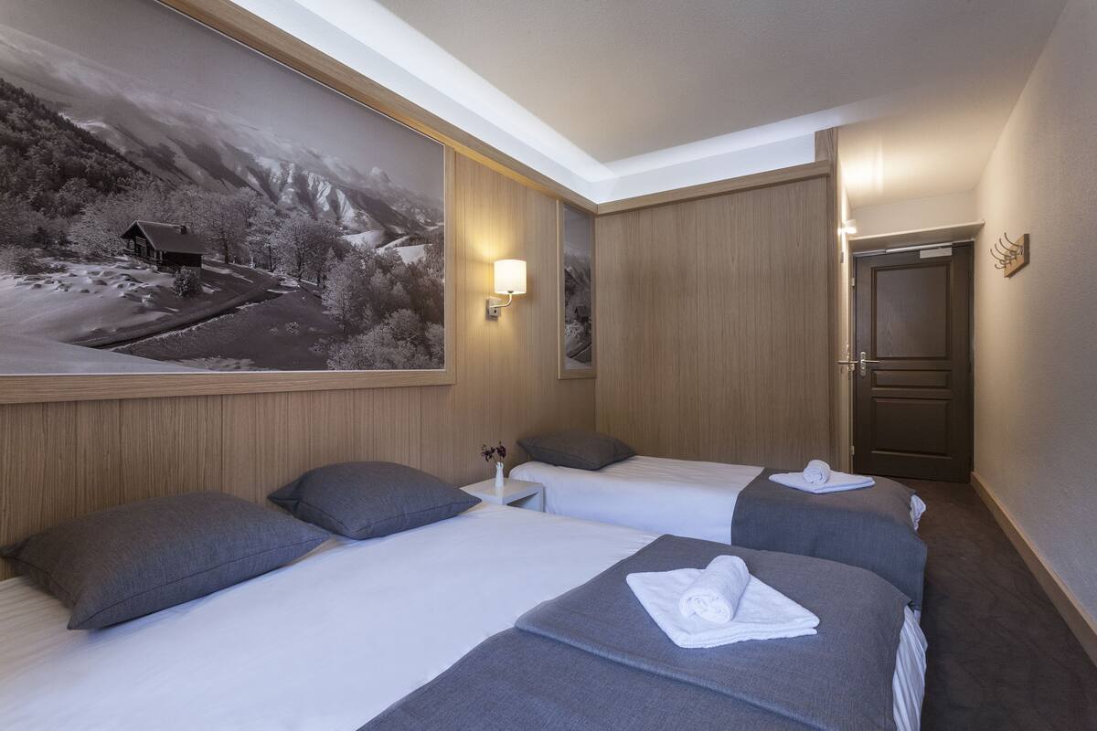 Habitación de hotel con dos camas, un cuadro grande enmarco que muestra un paisaje montañoso, una mesita de noche con una lámpara y una puerta de madera.