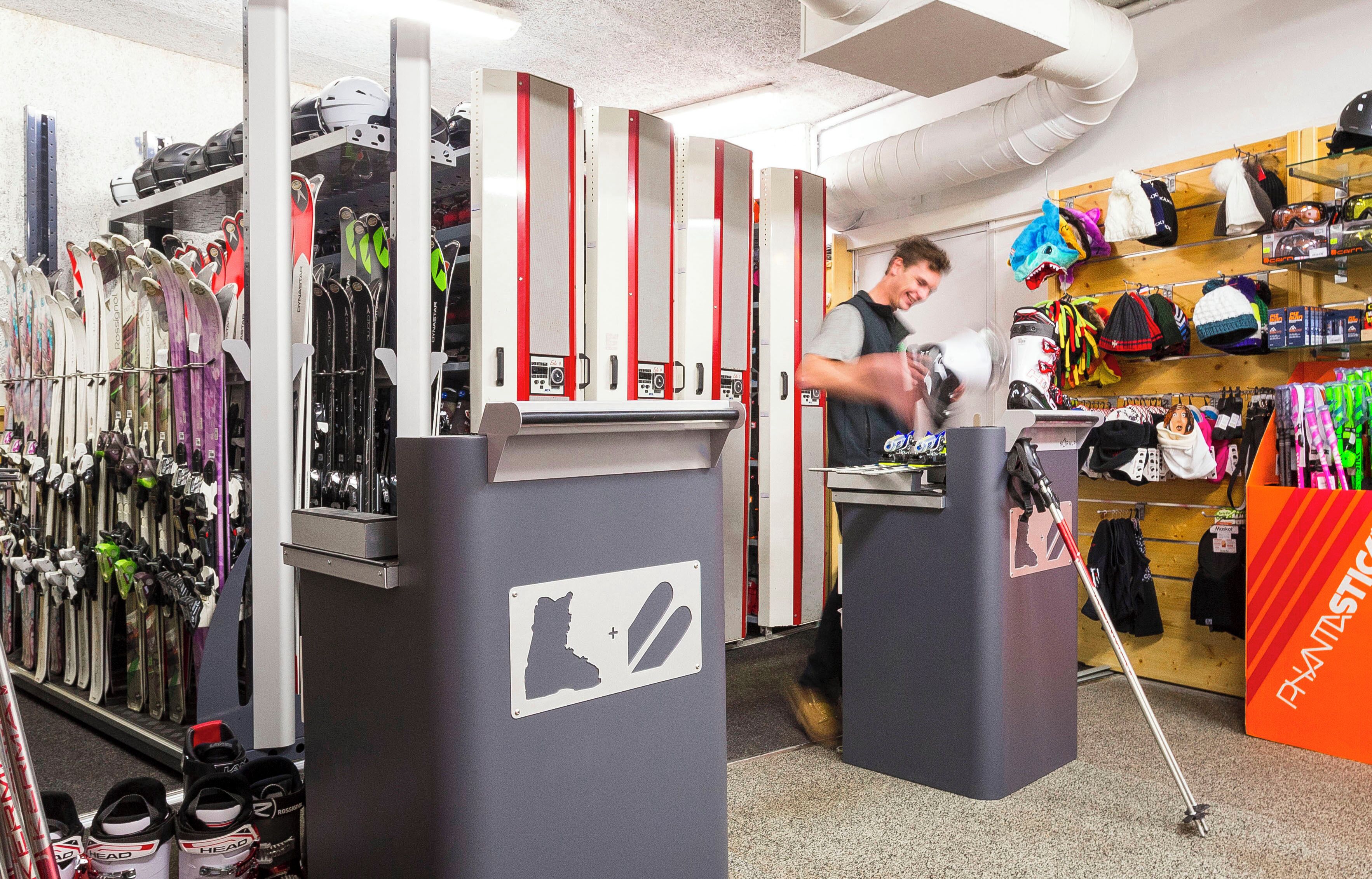 Tienda de equipo de esquí con estantes llenos de esquíes y tablas de snowboard, un mostrador con una persona atendiendo y una exhibición de ropa y accesorios para esquiar.