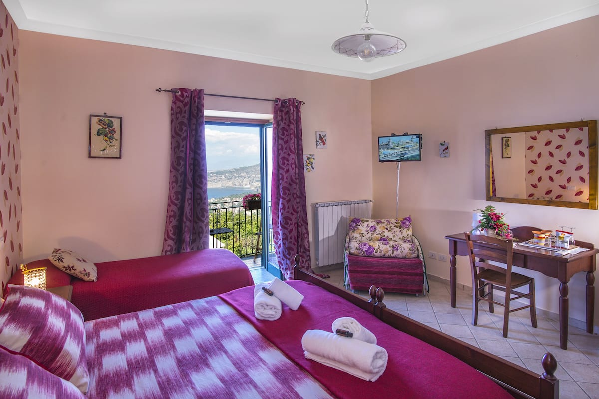 Ein Schlafzimmer mit zwei Betten, einem Schreibtisch und einem Stuhl. Es gibt ein Fenster mit Vorhängen, einen Fernseher an der Wand und Meerblick.