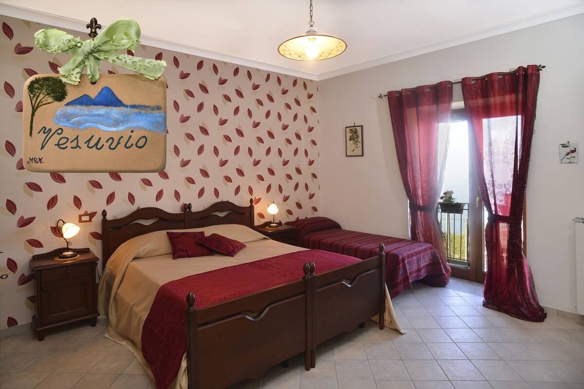Ein Schlafzimmer mit einem Bett, Nachttischen, einem Fenster mit Vorhängen sowie einer Wand mit Blättermotiv und einem Gemälde.