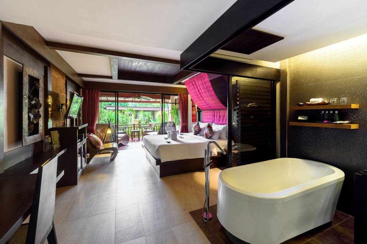 A modern hotel room with a large bed, a bathtub, a dining area, and a view of greenery outside.