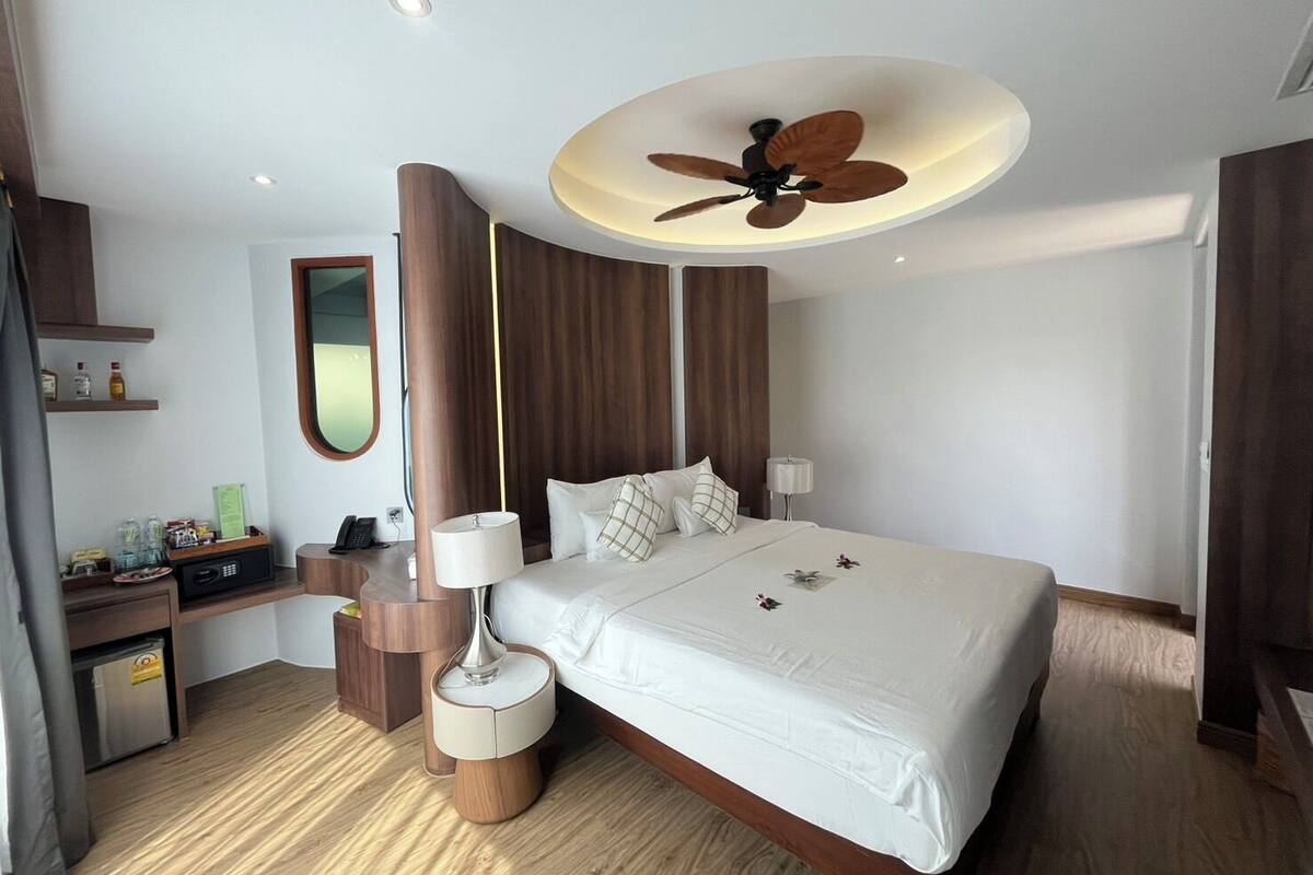 A modern hotel room with a large bed, a ceiling fan, a bedside table with a lamp, and a small desk with a telephone.