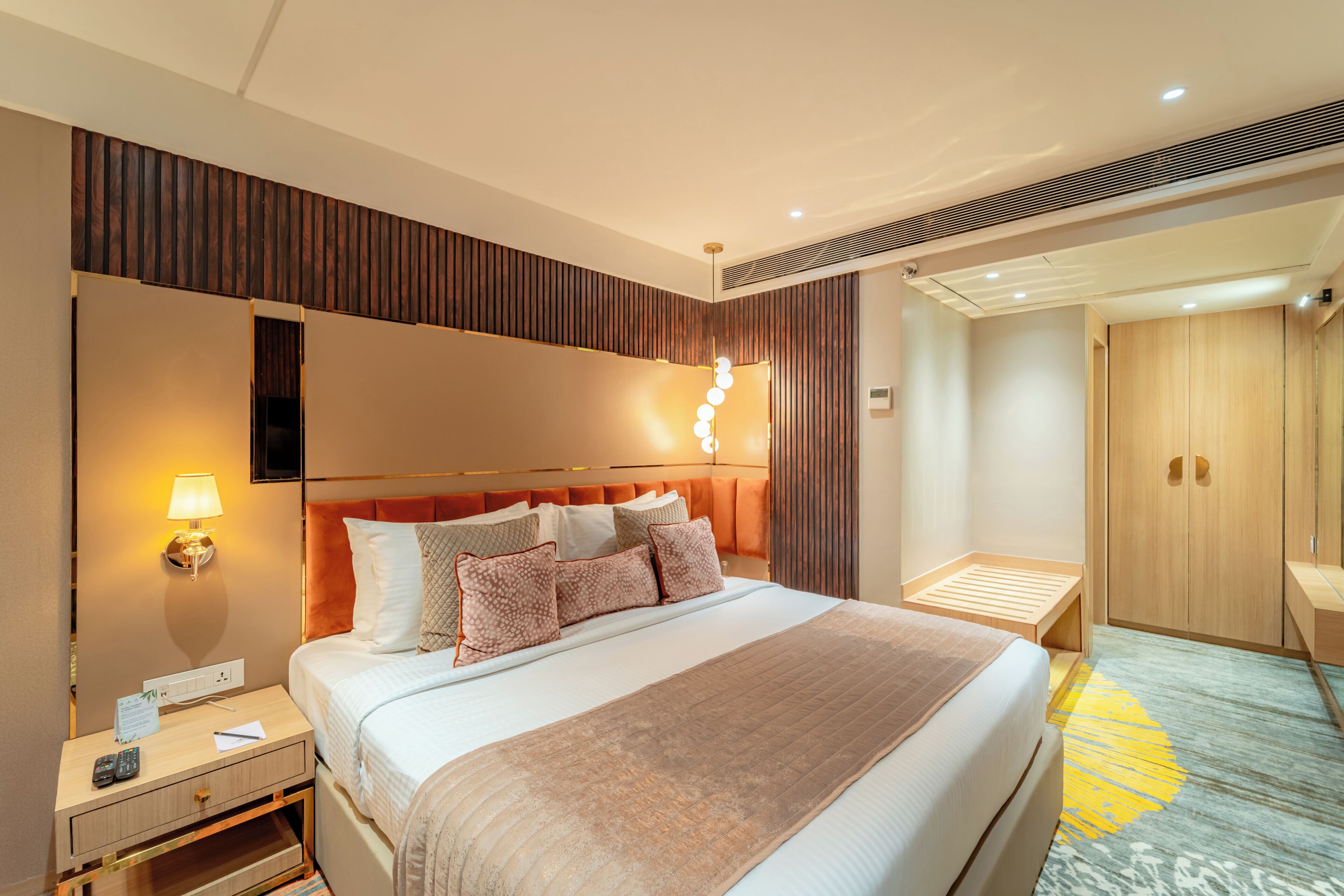 A modern hotel room with a large bed, a nightstand with a lamp, a wooden wardrobe, and a small seating area.