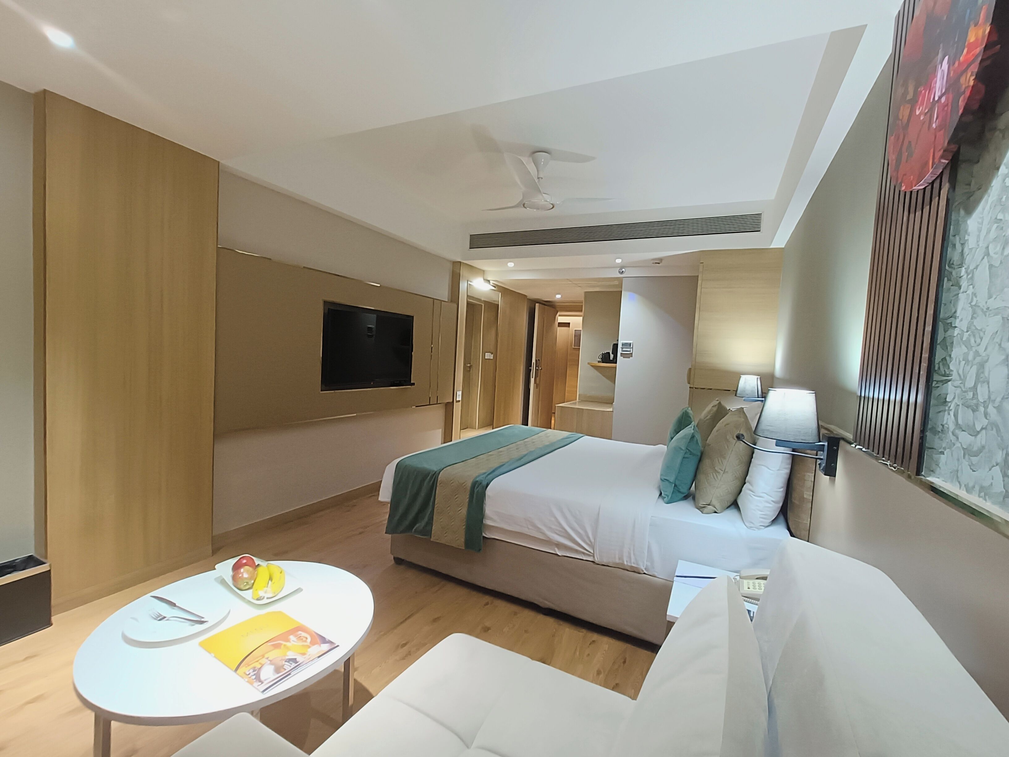 Une chambre d’hôtel avec un lit, un téléviseur fixé au mur, une petite table avec une assiette de fruits, un canapé et un ventilateur de plafond.