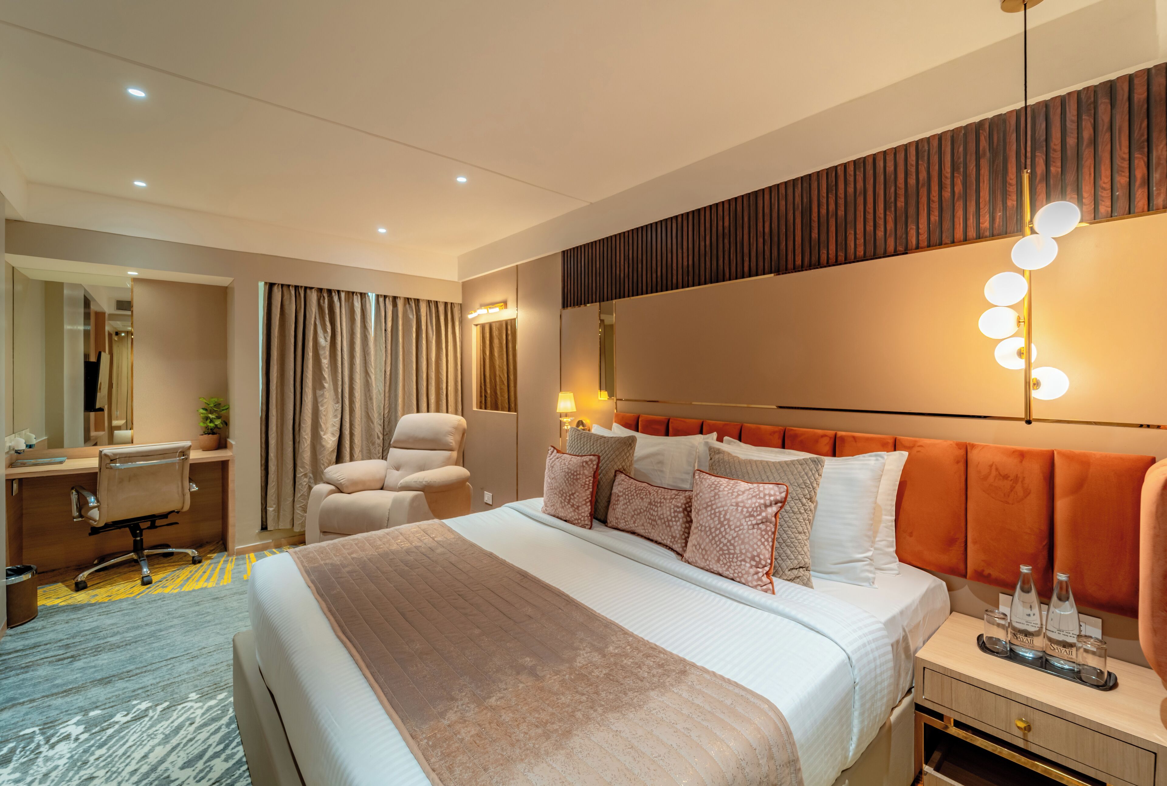 A modern hotel room with a large bed, a desk, a chair, a mirror, and a nightstand with a lamp.