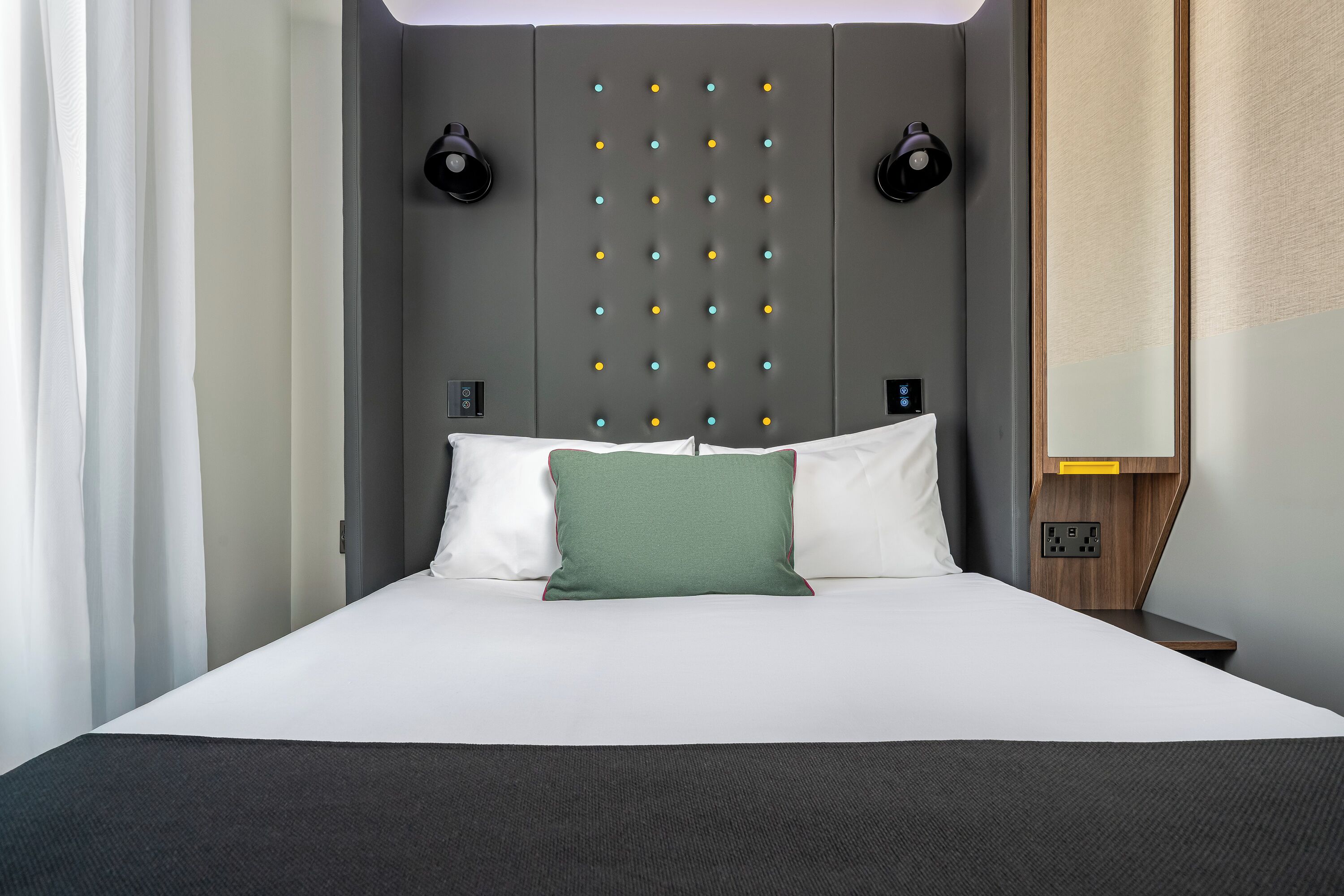 Une chambre d’hôtel moderne avec un grand lit, une tête de lit ornée d’une grille de lumières, des tables de chevet et une petite table de nuit avec une lampe.