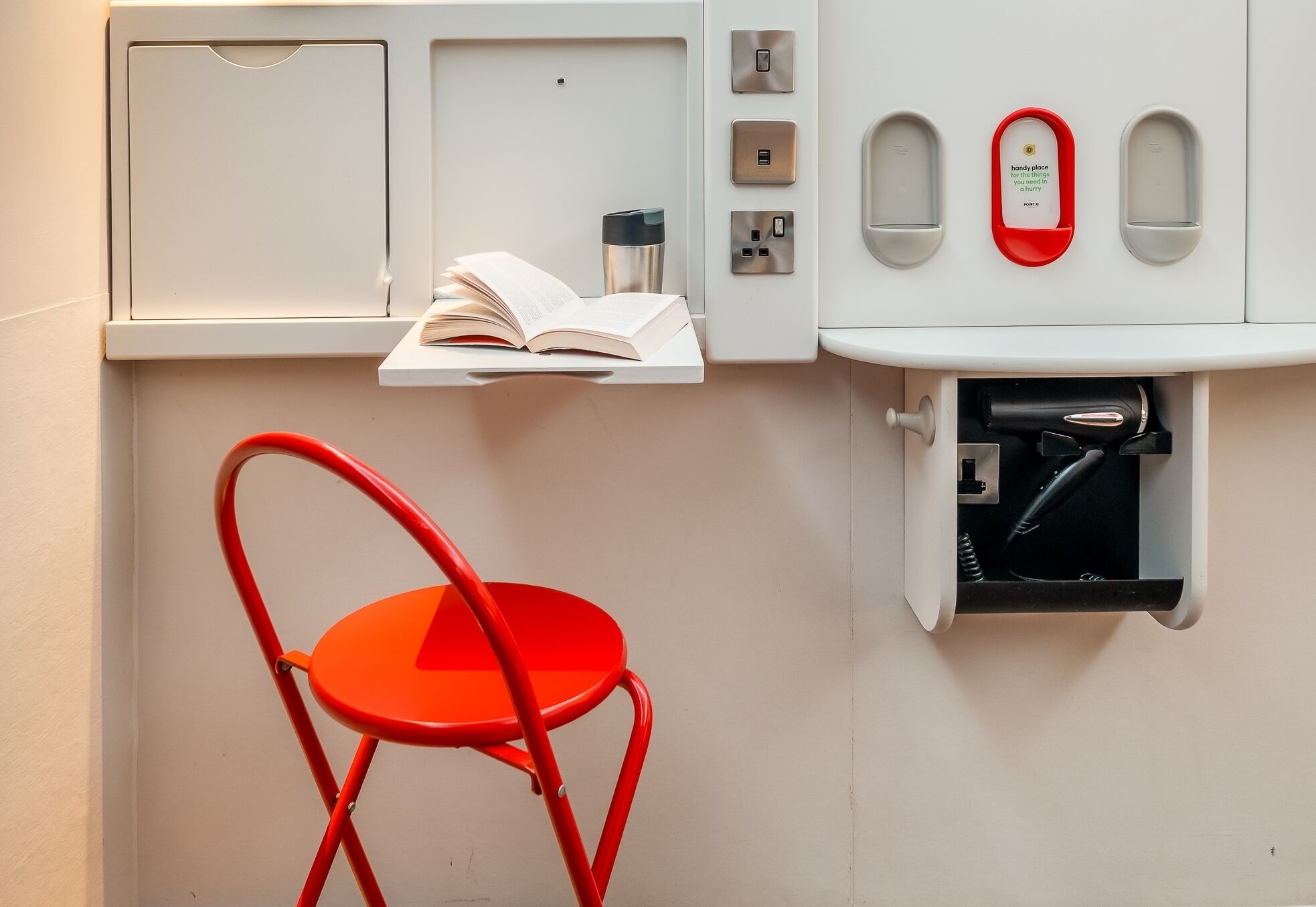 Une petite cuisine équipée, avec un plan de travail, une chaise rouge, un livre et un téléphone rouge réservé aux urgences.