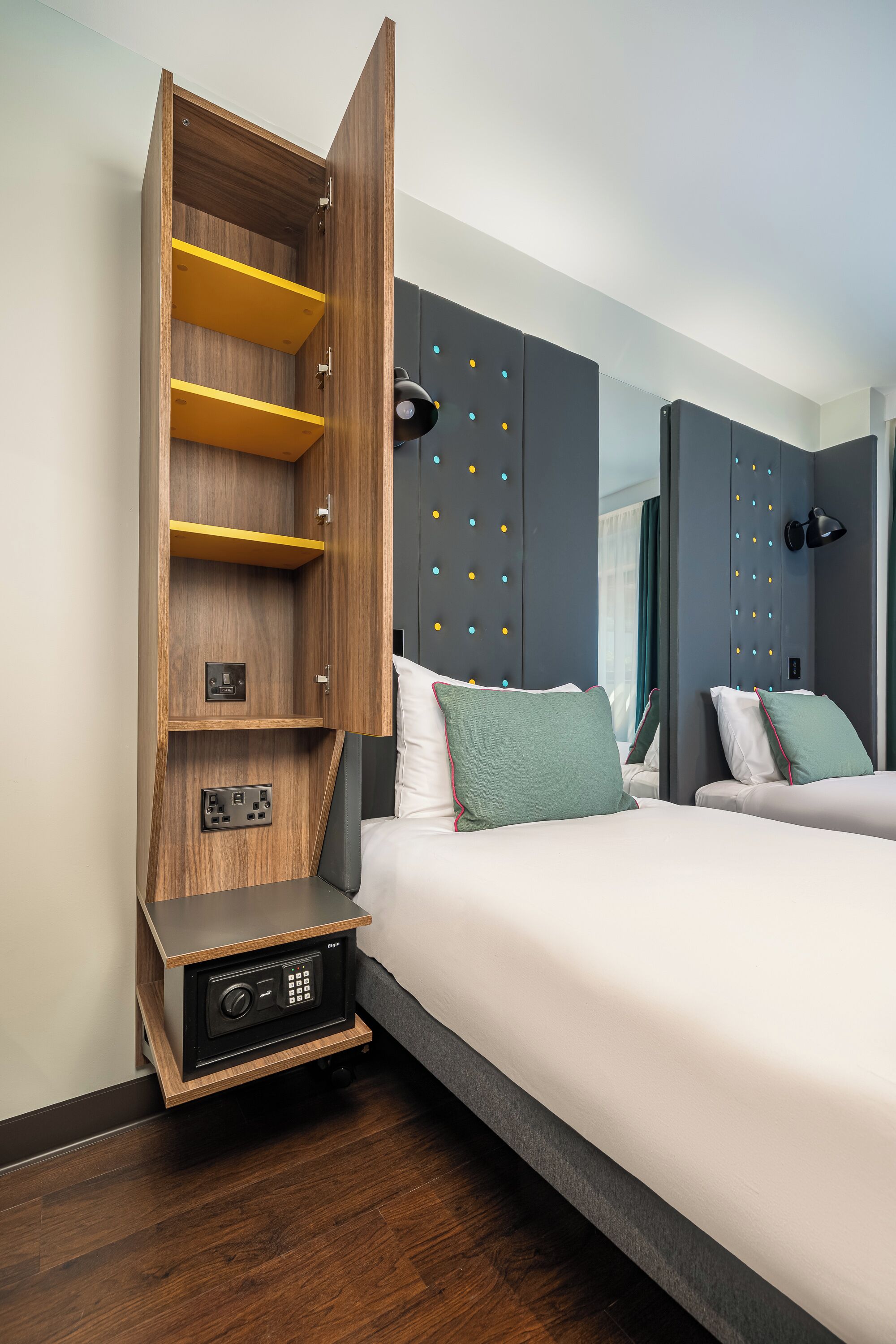 A hotel room with a built-in wooden shelf unit, a bedside table with a radio, and a bed with two pillows.