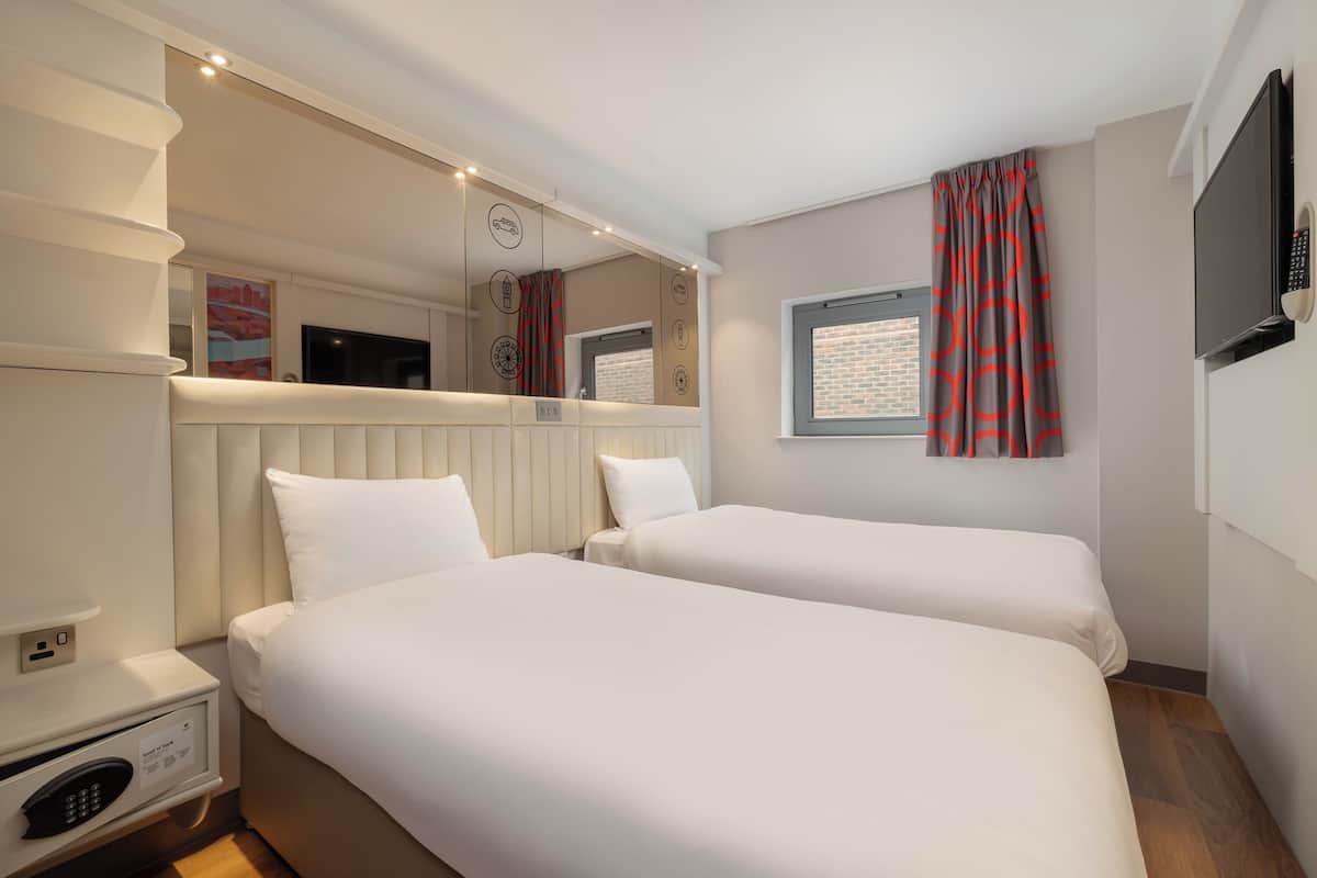 A hotel room with two beds, a TV, a mirror, a window with curtains, and a bedside table with a control panel.