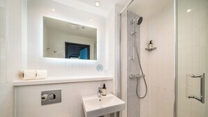 A modern bathroom with a white tiled wall, a rectangular mirror, a sink with a soap dispenser, and a shower area with a glass door. - Point A London Canary Wharf  (London)