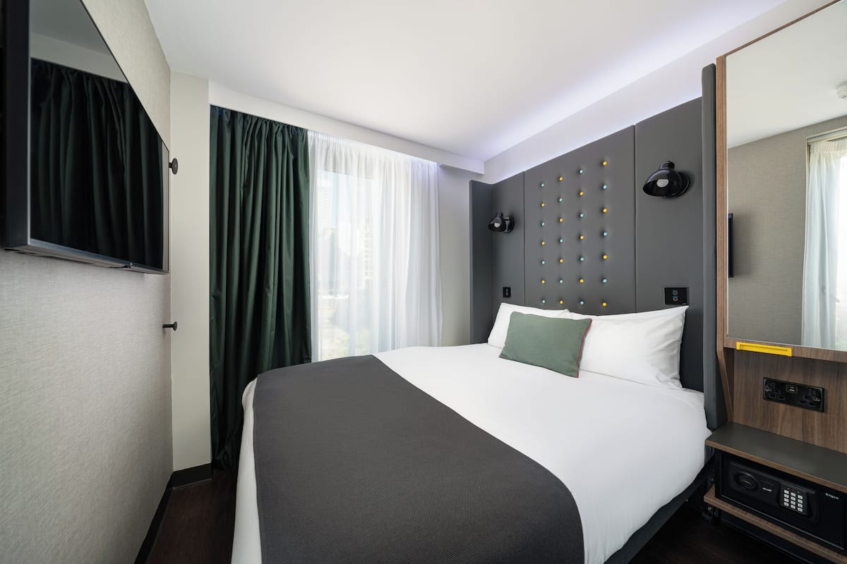 A modern hotel room with a large bed, a flat-screen TV mounted on the wall, a bedside table with a lamp, and a large mirror.