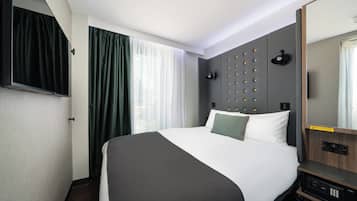 A modern hotel room with a large bed, a flat-screen TV mounted on the wall, a bedside table with a lamp, and a large mirror.