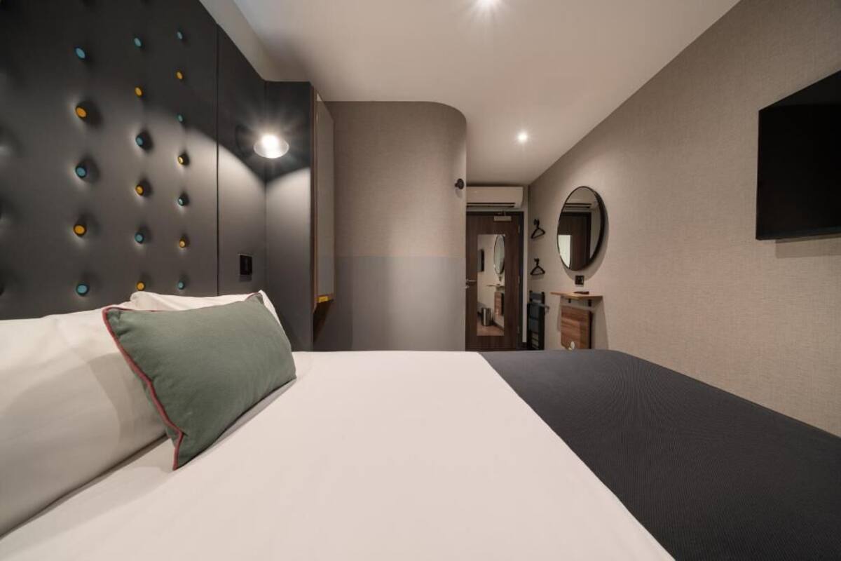 A modern hotel room with a large bed, a built-in headboard with circular lights, a flat-screen TV, and a mirror on the wall.