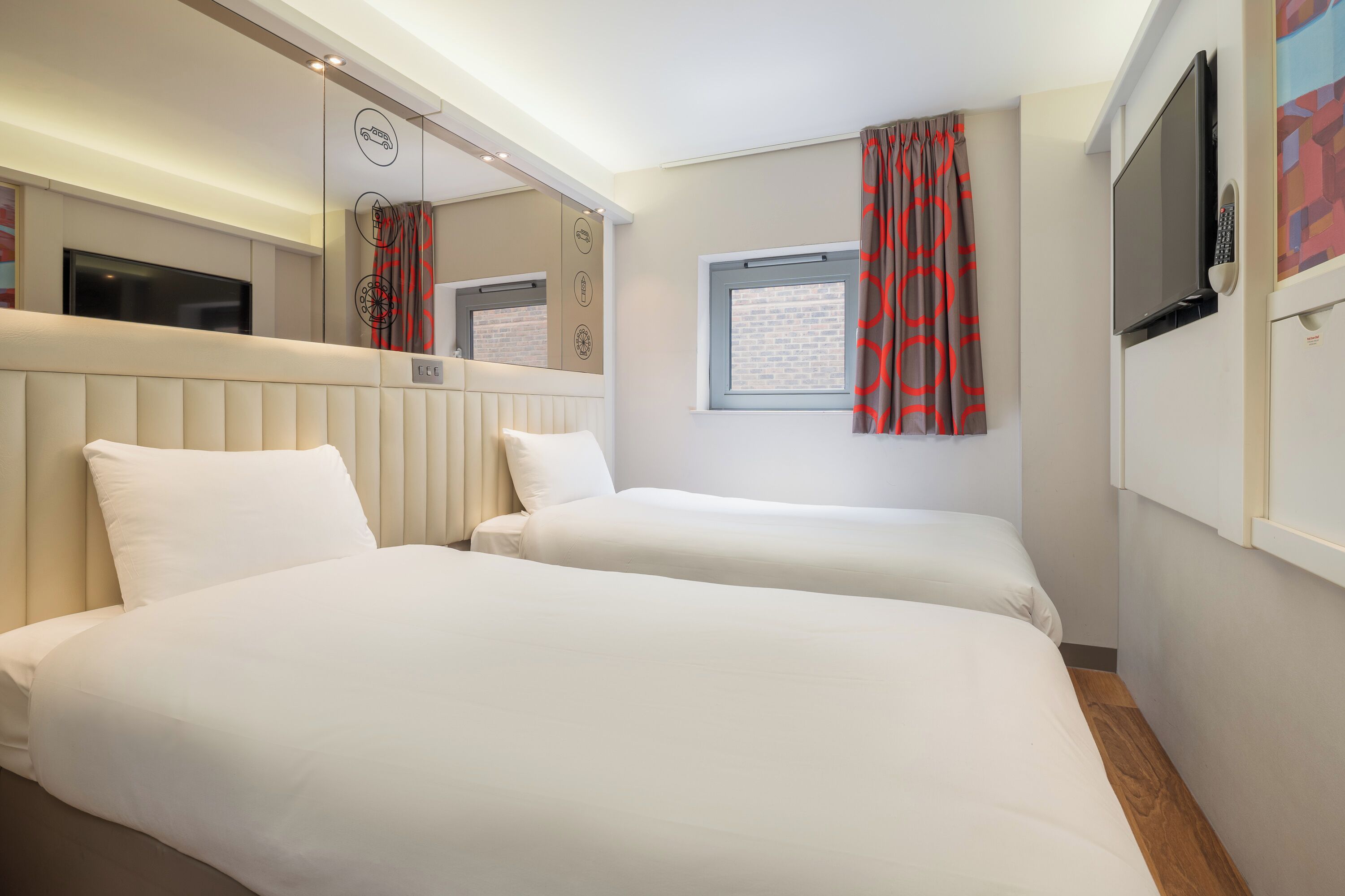 Habitación de hotel con dos camas, televisión, ventana con cortinas y televisión instalada en la pared.