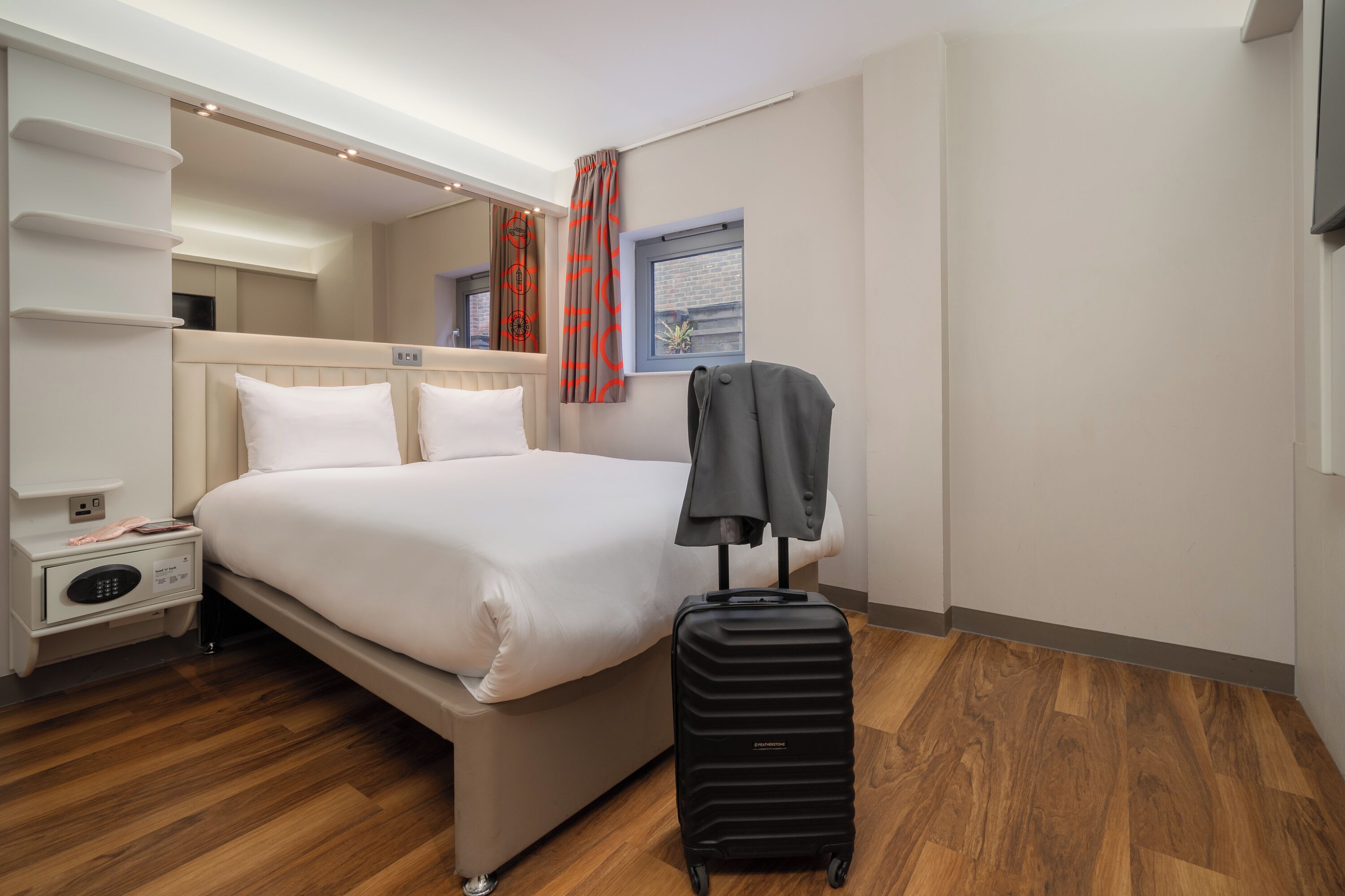 A hotel room with a bed, a suitcase, a nightstand with a phone, and a window with curtains.