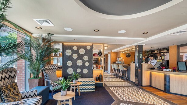 A modern hotel lobby with a reception desk, seating area, and a view of the kitchen. - Point A London Canary Wharf  (London)
