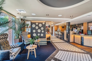 Un hall d’hôtel moderne avec un comptoir d’accueil, un espace salon et une vue sur la cuisine.