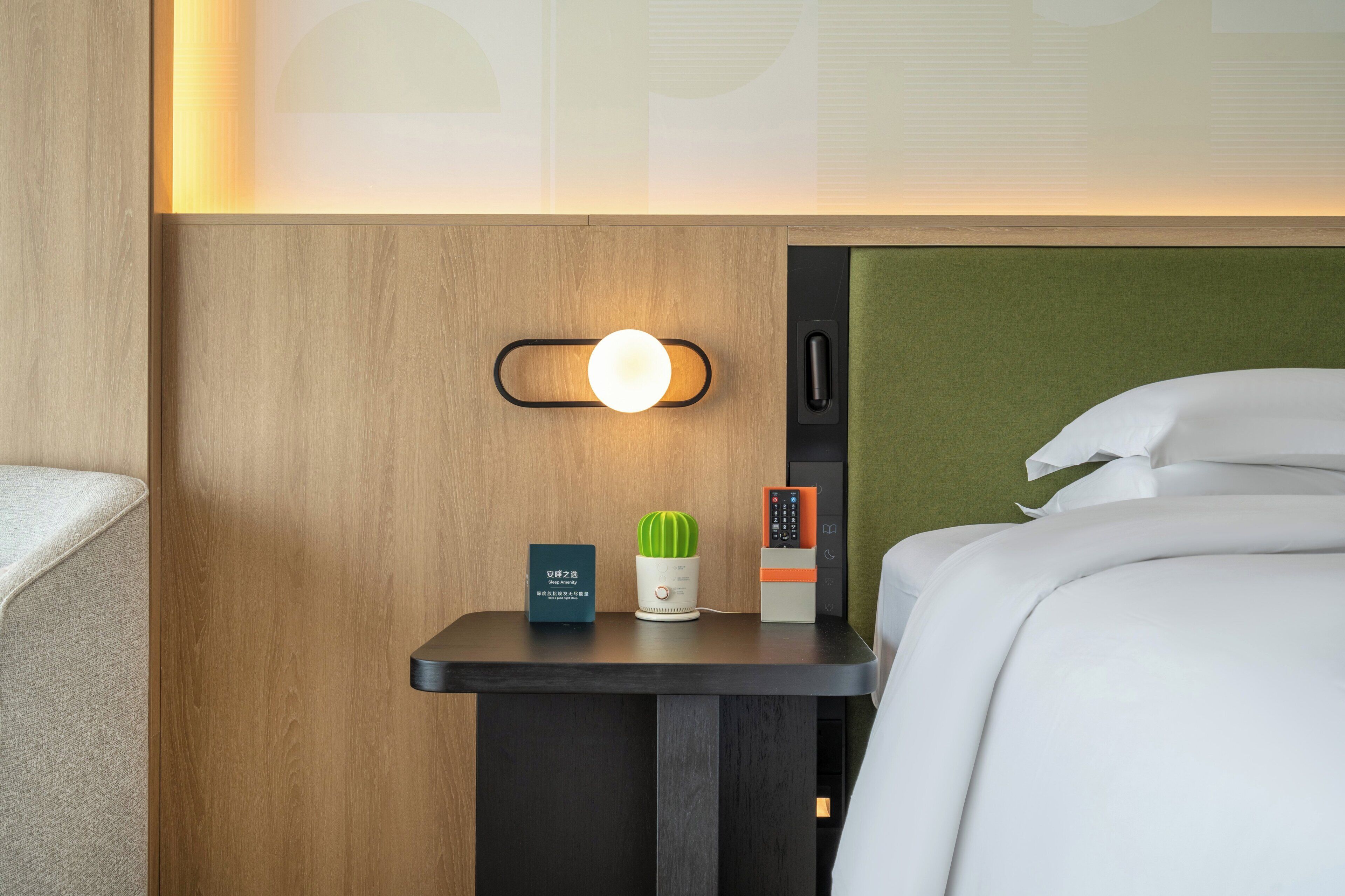 Een hotelkamer met een net opgemaakt bed, een nachtkastje met een lamp, een afstandsbediening en een kleine potplant.