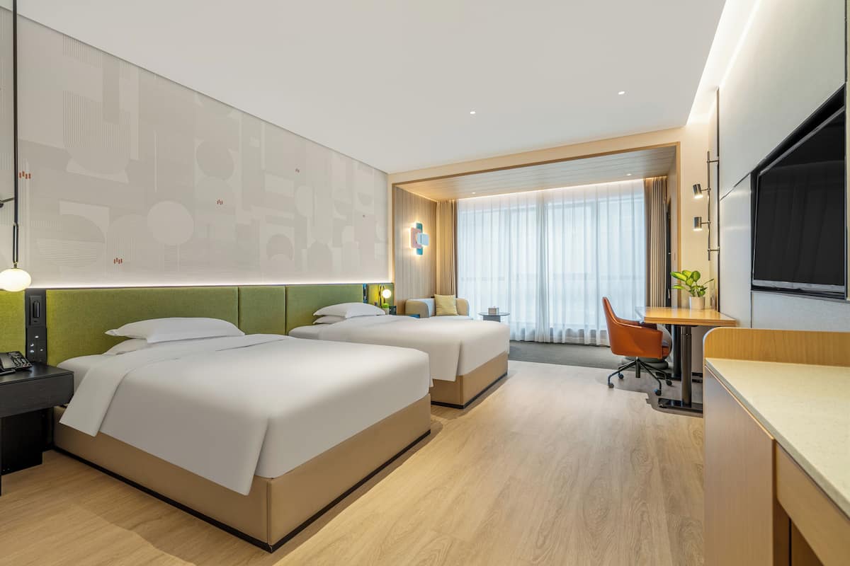 A hotel room with two beds, a desk, a chair, a TV, and a large window with curtains.