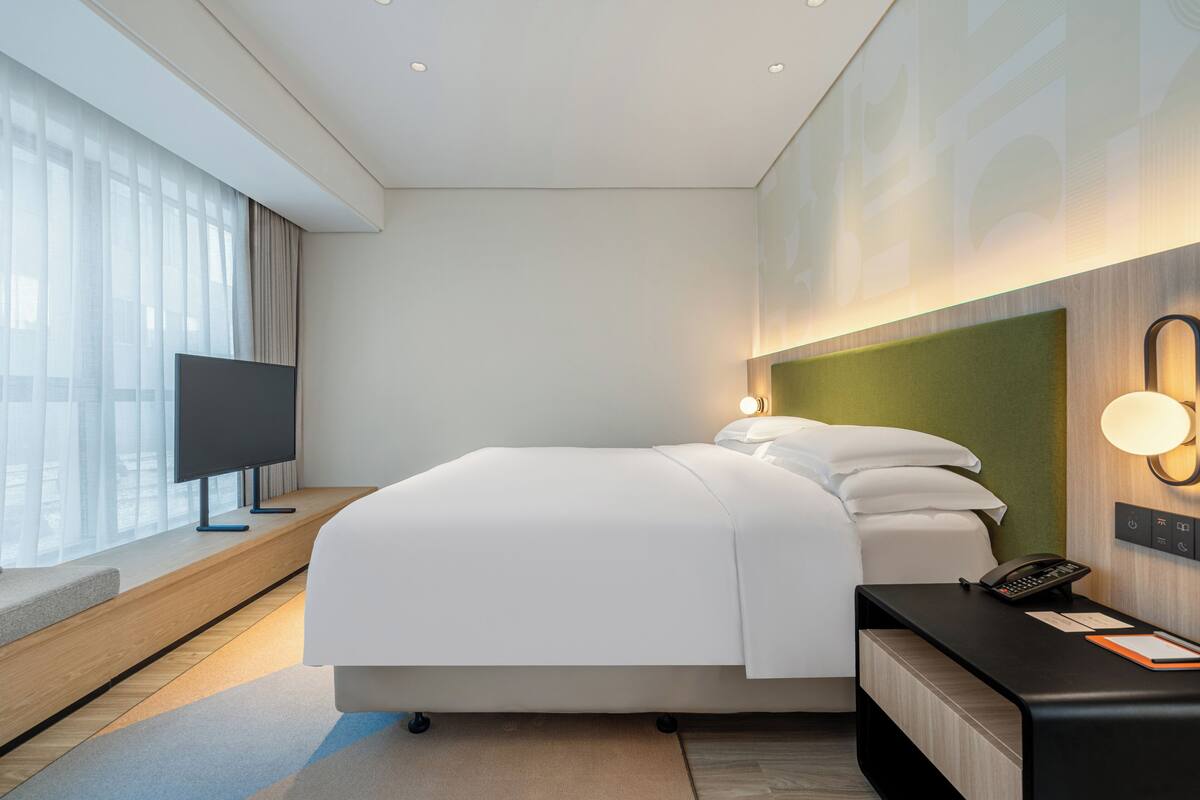 A modern hotel room with a large bed, a flat-screen TV, a bedside table with a phone, and a view of the city through large windows.