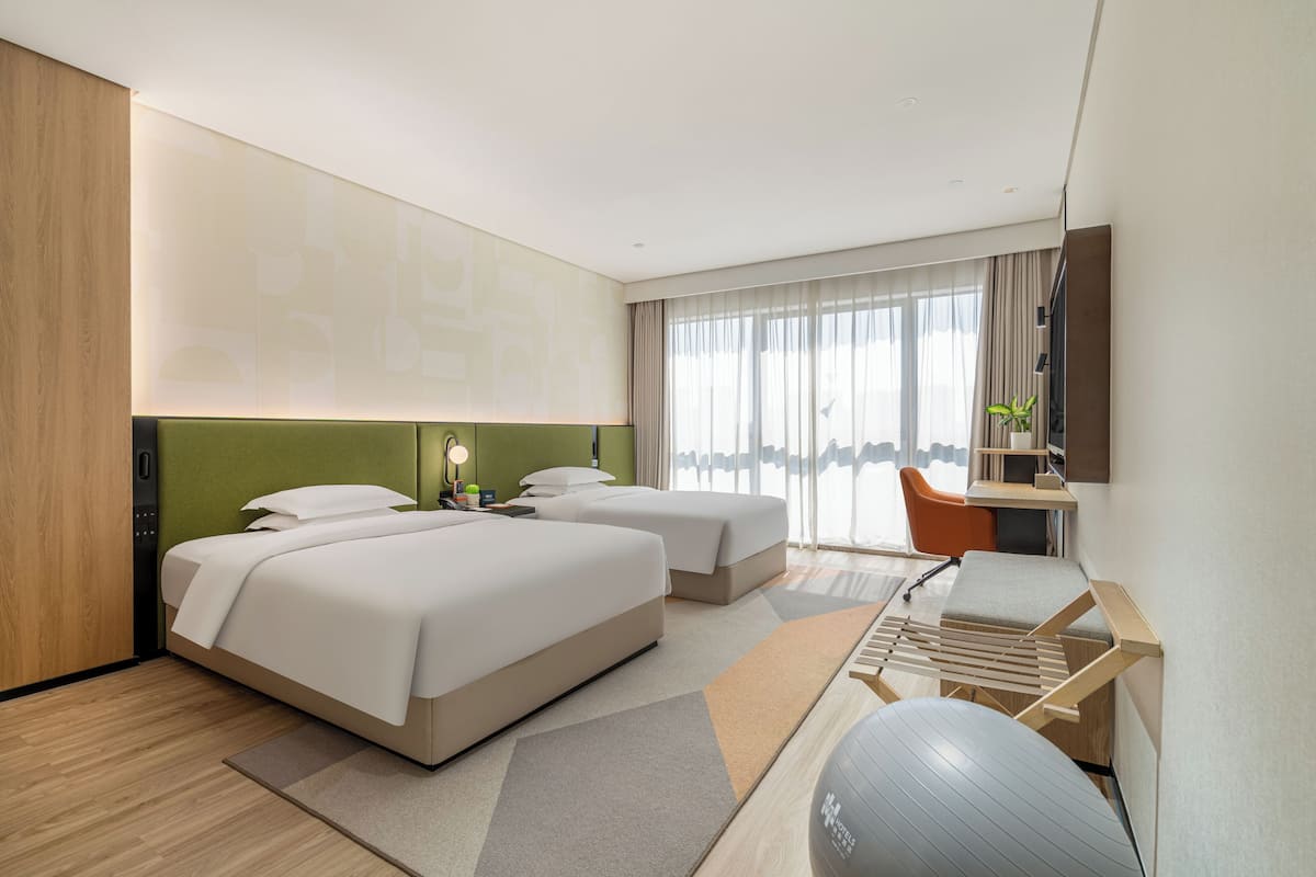 A modern hotel room with two beds, a desk, a chair, a treadmill, and a large window with curtains.