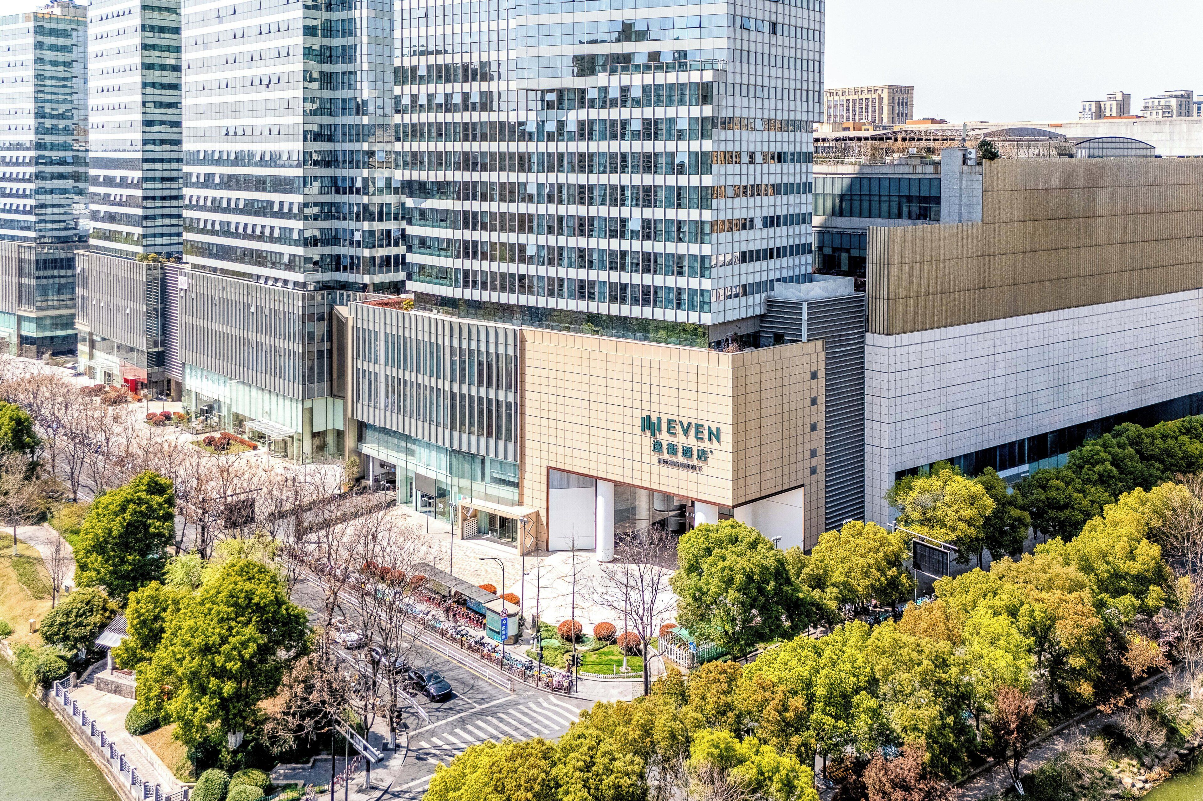 Un edificio moderno con fachada de vidrio, un cartel que dice 'WEVEN', y un parque circundante con árboles.