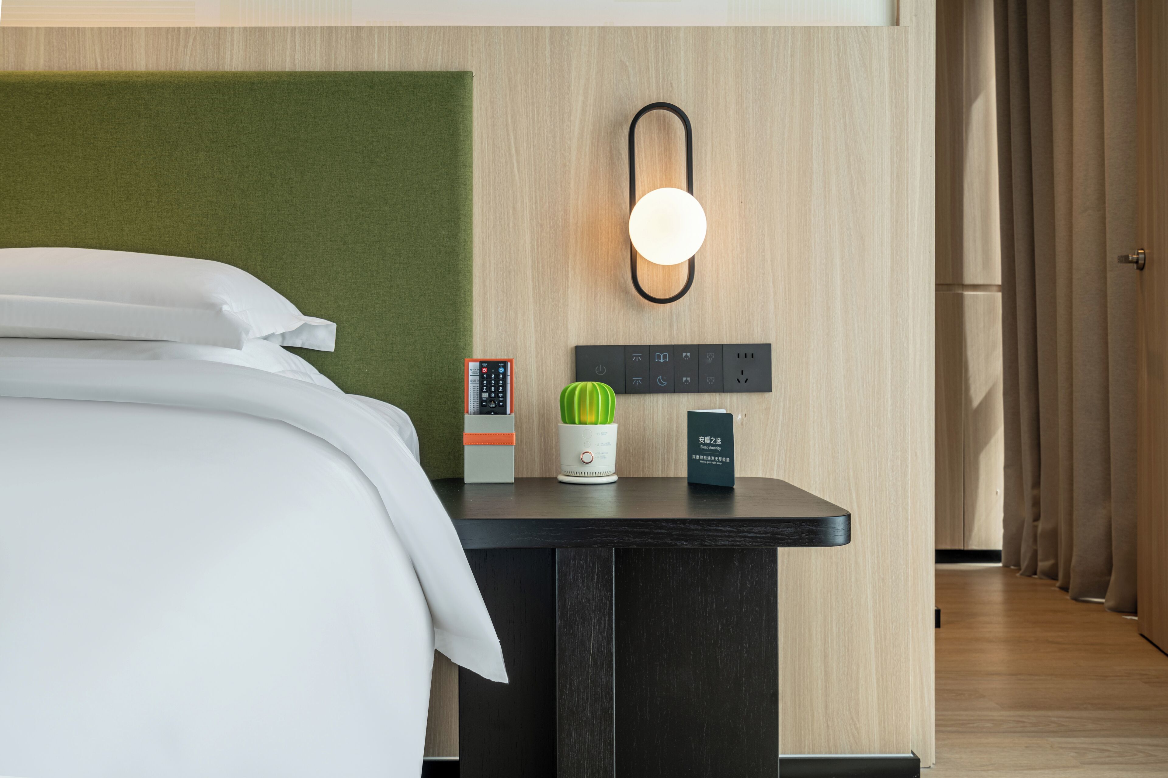 Une chambre d’hôtel avec un lit, une table de chevet avec une lampe, un thermostat, une plante et une porte.
