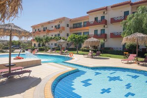 A swimming pool with lounge chairs and umbrellas, surrounded by a building with balconies. - Portokali Apartments (Hersonissos)