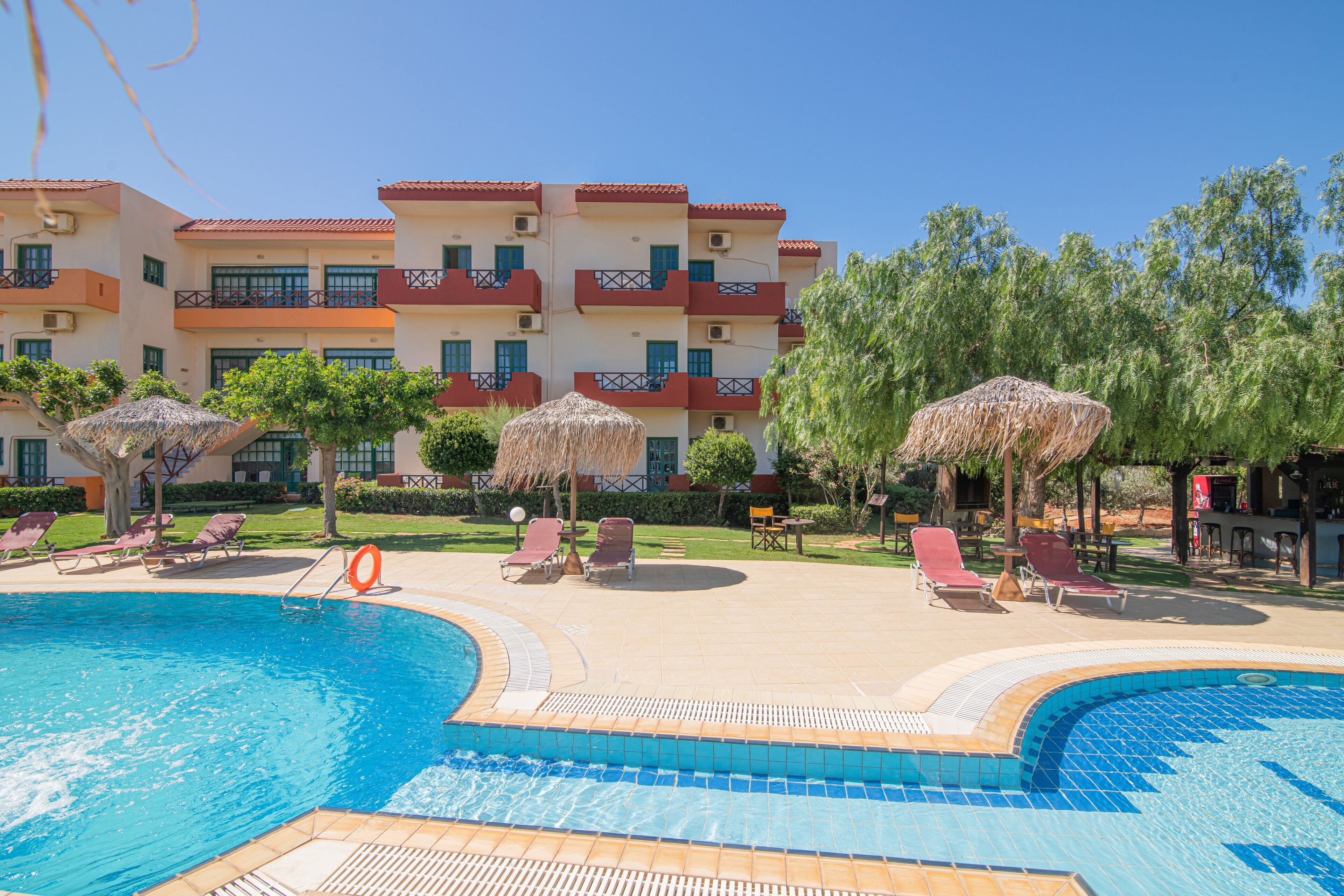 Zona de piscina de hotel con múltiples sillones de descanso, un puesto de salvavidas y un edificio con balcones.