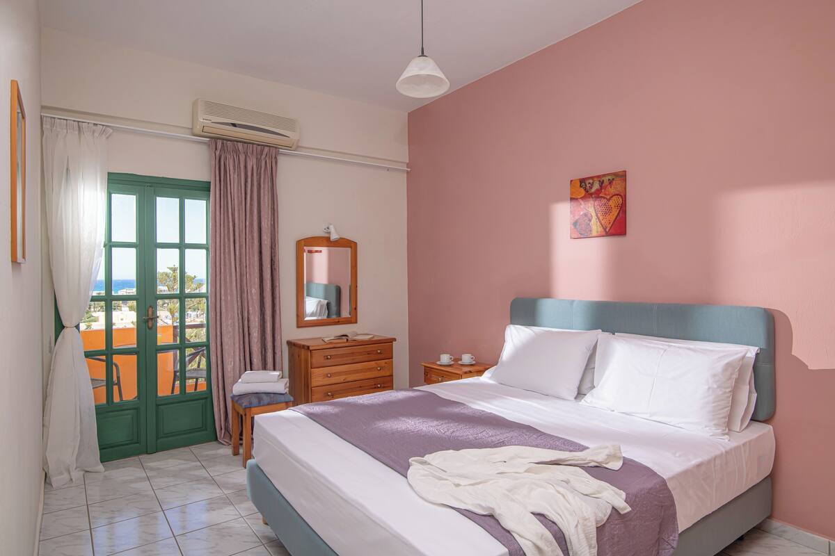 Une chambre à coucher avec un lit, une commode, un miroir, une fenêtre avec des rideaux et un tableau accroché au mur.