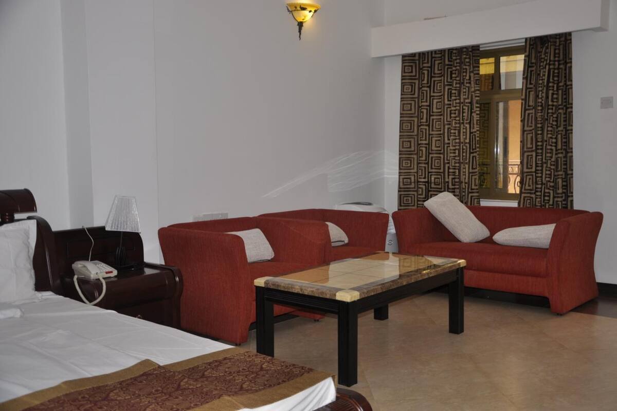 Une chambre d’hôtel avec deux canapés rouges, une table basse en bois, un lit avec du linge de maison blanc, un téléphone et un rideau à motifs.