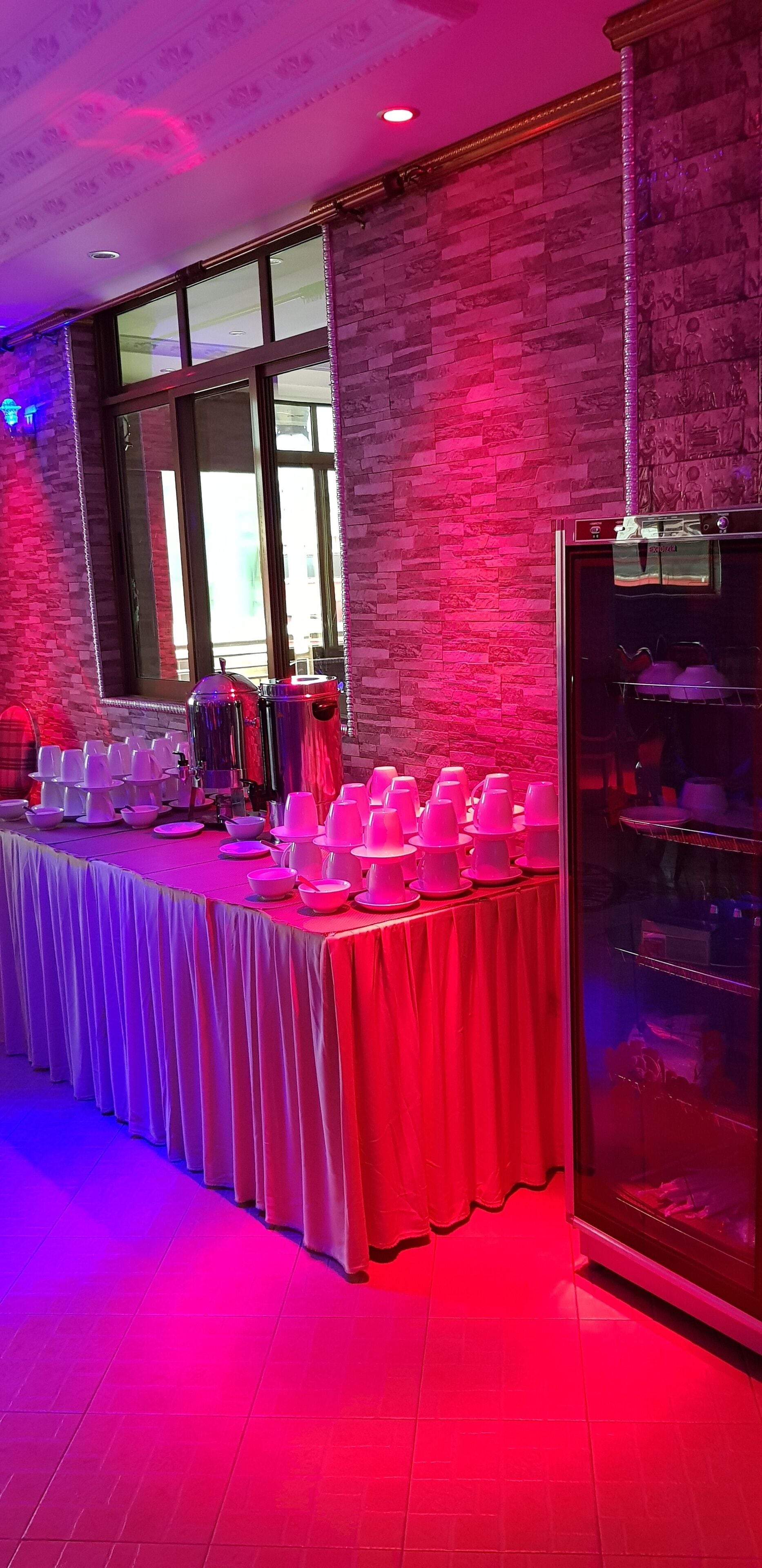Un salón de banquetes con una larga mesa cubierta con un mantel blanco, rodeada de tazas y cuencos rosa y blancos, y una vitrina de vidrio con diversos objetos.