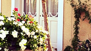 Un edificio con una puerta blanca, una palmera y diversas plantas con flores.