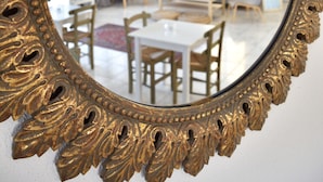 Un espejo con marco dorado ornamentado refleja una sala de estar moderna con comedor, sofá y mesa de centro.