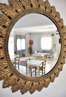 Un espejo con marco dorado ornamentado refleja una sala de estar moderna con comedor, sofá y mesa de centro.