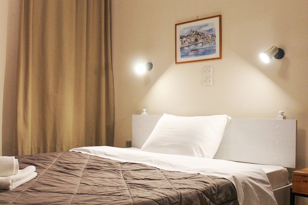 Een net opgemaakt bed met een bruine dekbedovertrek en witte kussens, een ingelijst beeld aan de muur, en een nachtkastje met een lamp.