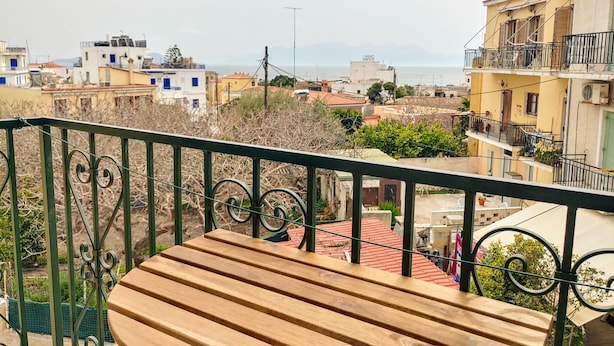 Un balcón con banca de madera, con vista a una zona residencial con edificios y una lejana vista al mar.