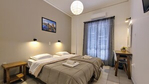 Habitación de hotel con dos camas, un escritorio, una silla, una mesita pequeña, una planta en maceta y un ventanal con cortinas.
