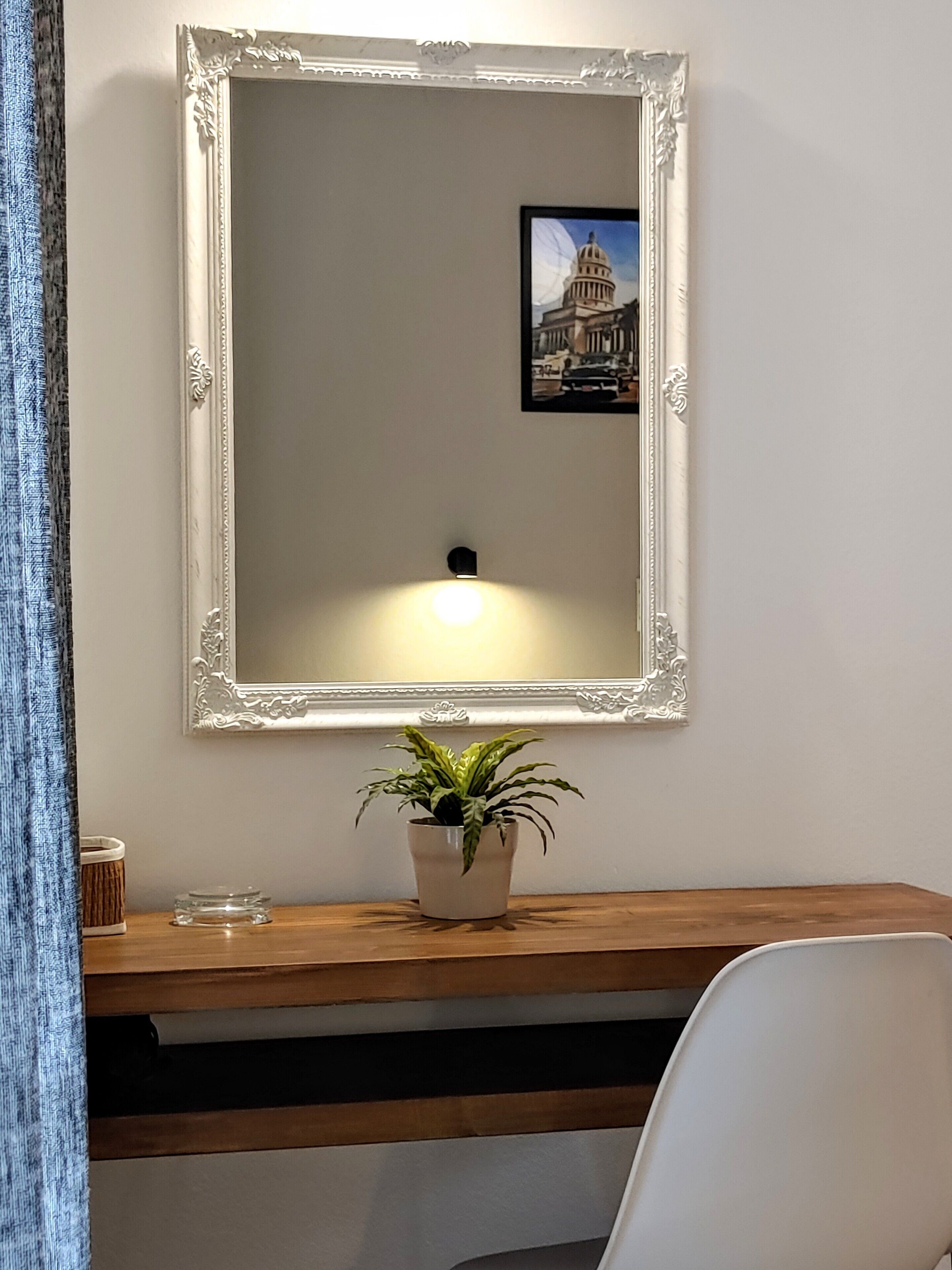 A mirror with an ornate frame, a desk with a potted plant, and a framed picture of a building on the wall.