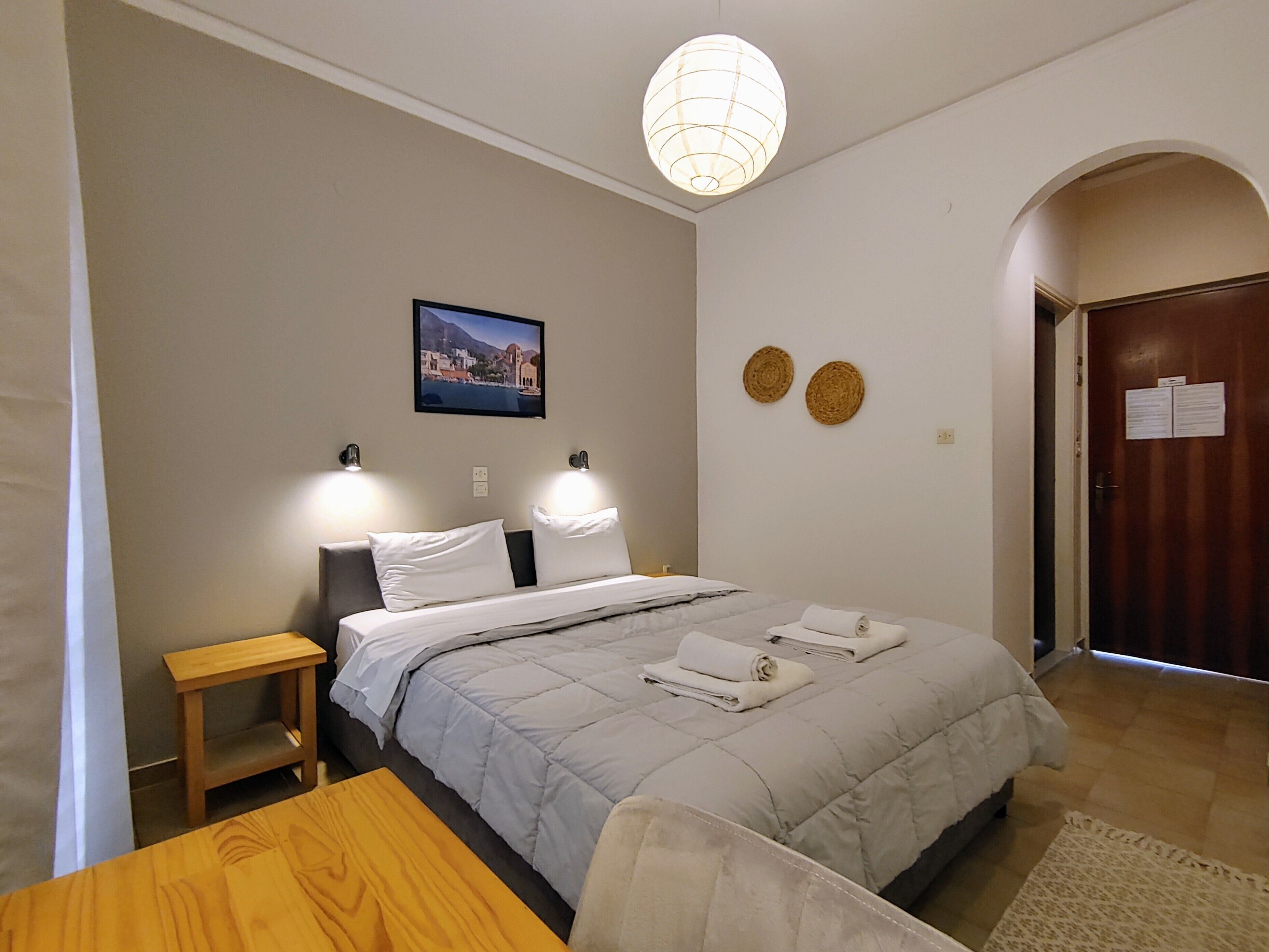 Un dormitorio ordenado con cama, mesitas de noche, una mesa de madera, una lámpara colgante y una puerta con un letrero.