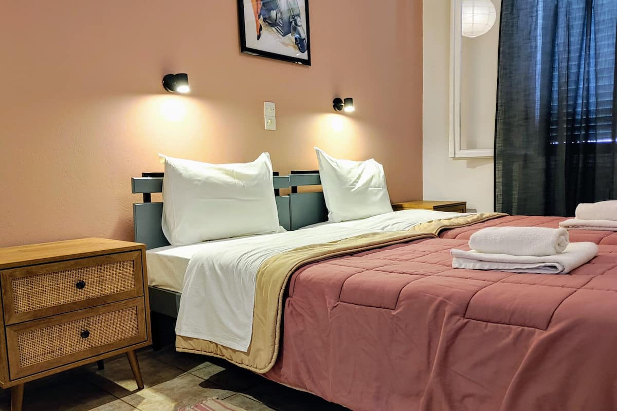 Een netjes opgemaakt bed met een roze dekbedovertrek en witte kussens, een nachtkastje met een lamp, en een gestreept vloerkleed.