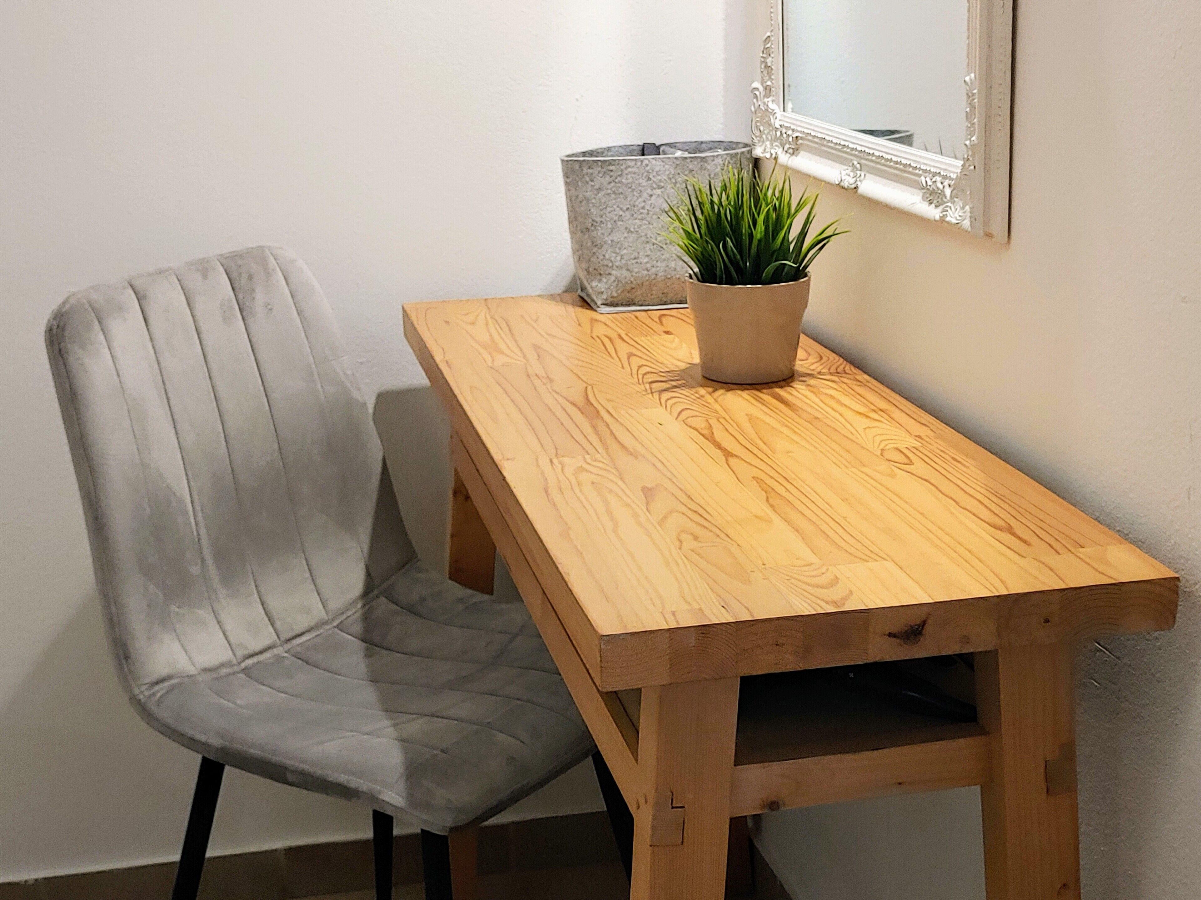 Una mesa de madera con una planta en maceta, una silla tapizada en gris y una pared blanca con un espejo.