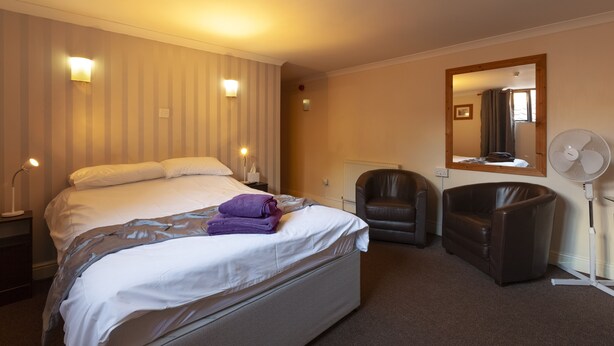 A hotel room with a large bed, a chair, a standing fan, and a mirror on the wall. - The William IV (Norwich)