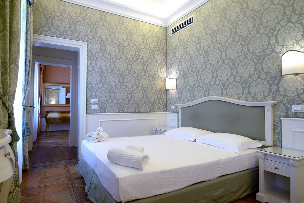A neatly made bed with white linens and towels, a bedside table with a lamp, and a patterned wallpaper.