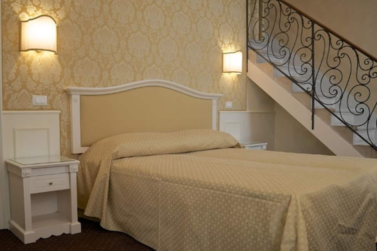 Un lit bien fait, avec une tête de lit, deux tables de chevet et des lampes fixées au mur. Un escalier avec une rampe en fer forgé est visible sur la droite.
