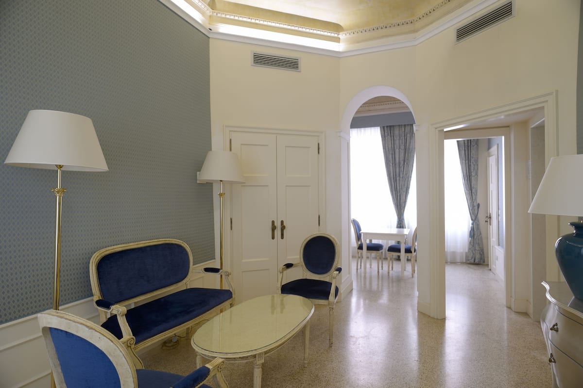 Une chambre d’hôtel avec un coin salon, une table et une lampe. On y trouve une porte et un arc en plein cintre qui mène à une autre pièce.