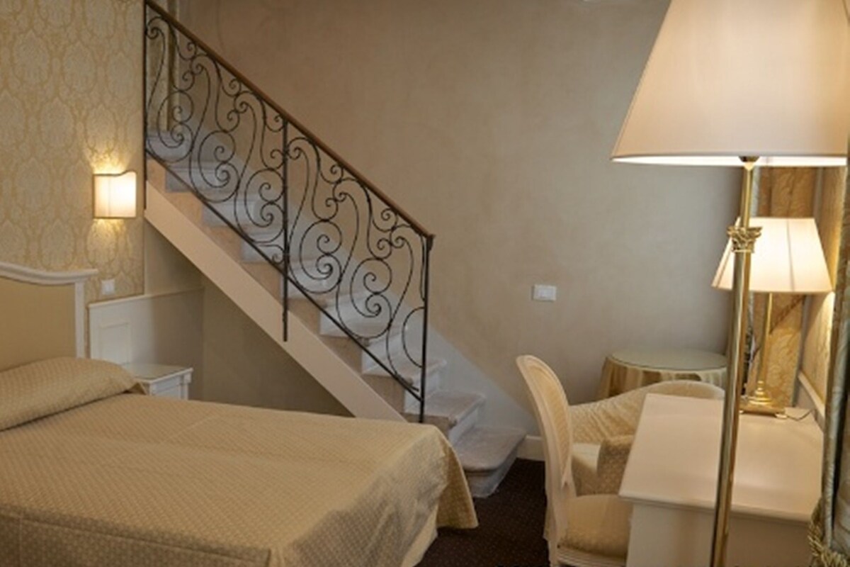 A hotel room with a bed, a chair, a desk, a lamp, and a staircase with a decorative railing.