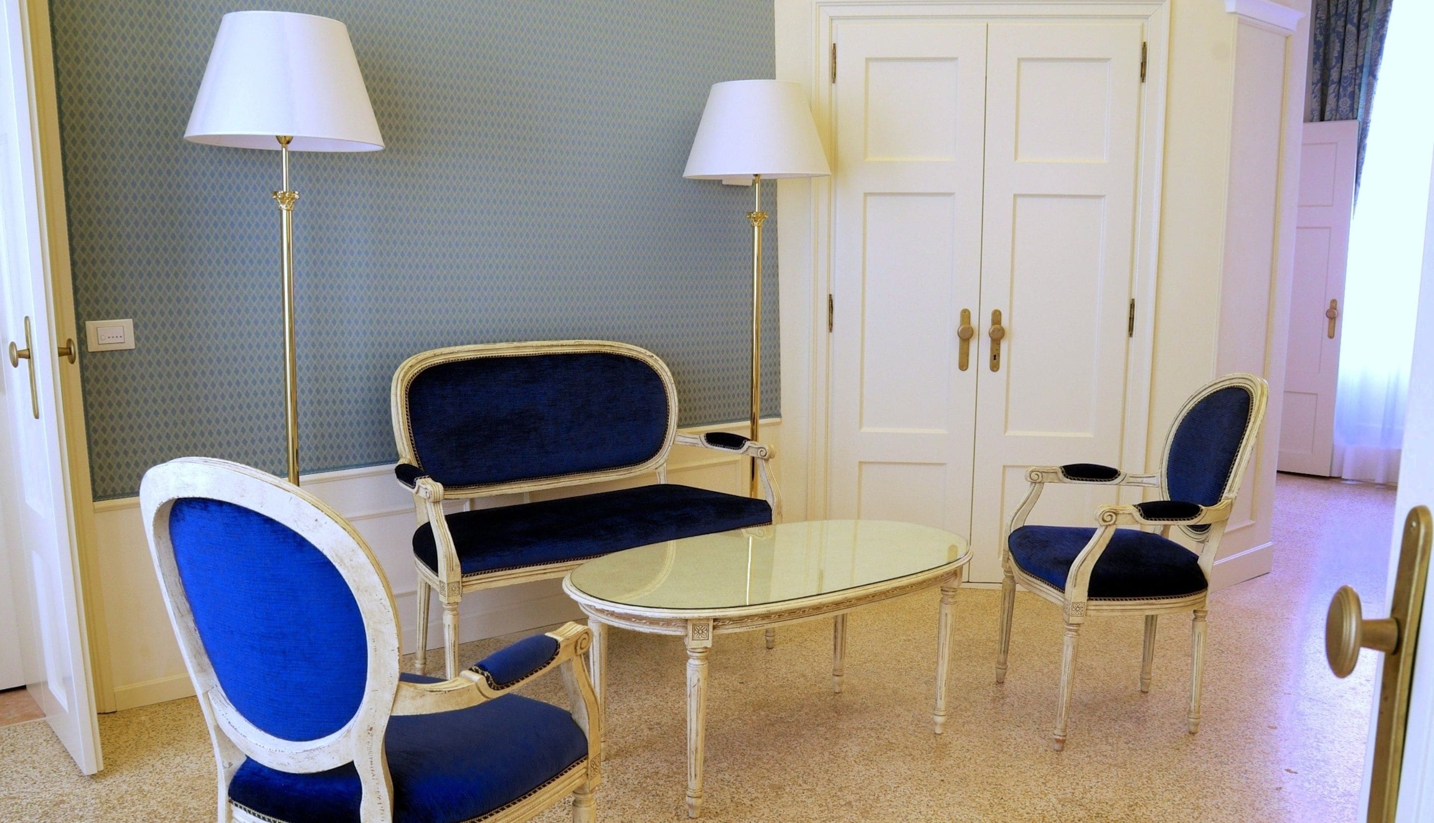 Une pièce comprenant un canapé bleu recouvert de tissu, deux fauteuils, une petite table ronde et deux lampadaires. On y trouve une porte blanche avec une poignée et un interrupteur sur le mur.